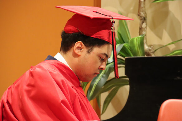 A student wearing a graduation gown and hat with a gold "24" on the tassel plays the piano