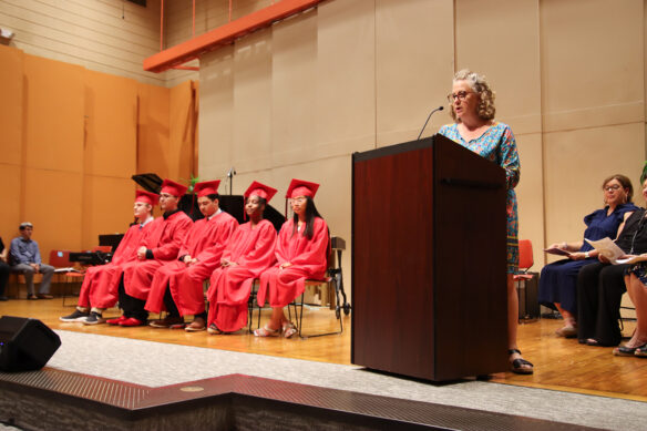 A woman talks into a microphone on a podium as five students in graduation gowns sit in chairs to her right