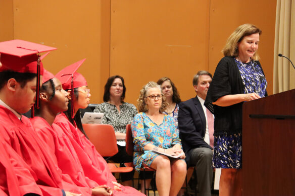 A woman talks into a mic on a podium while people sit behind her and students sit to the right of her in graduation gowns