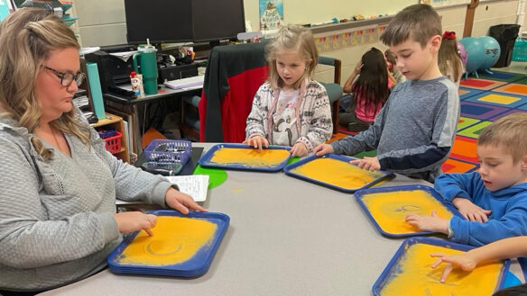 A teacher sits at a desk with kids who are all drawing in sand