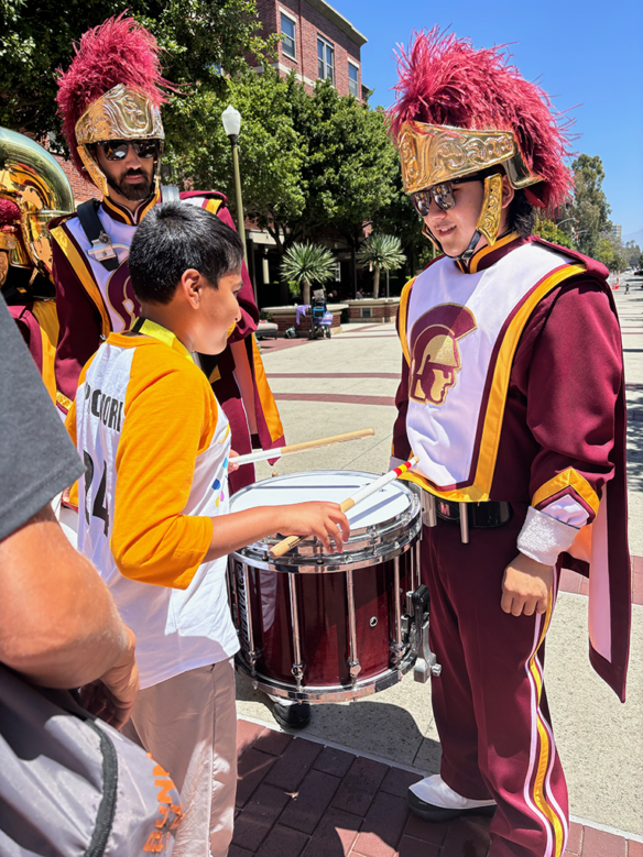 Krishna Malhotra stands outside in front of a University of Southern California drummer, holding onto drumsticks and getting ready to play the student's drum.