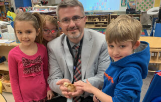 Robbie Fletcher poses with kids in a classroom, holding a baby chicken