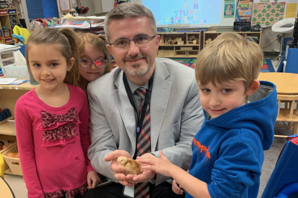 Robbie Fletcher poses with kids in a classroom, holding a baby chicken