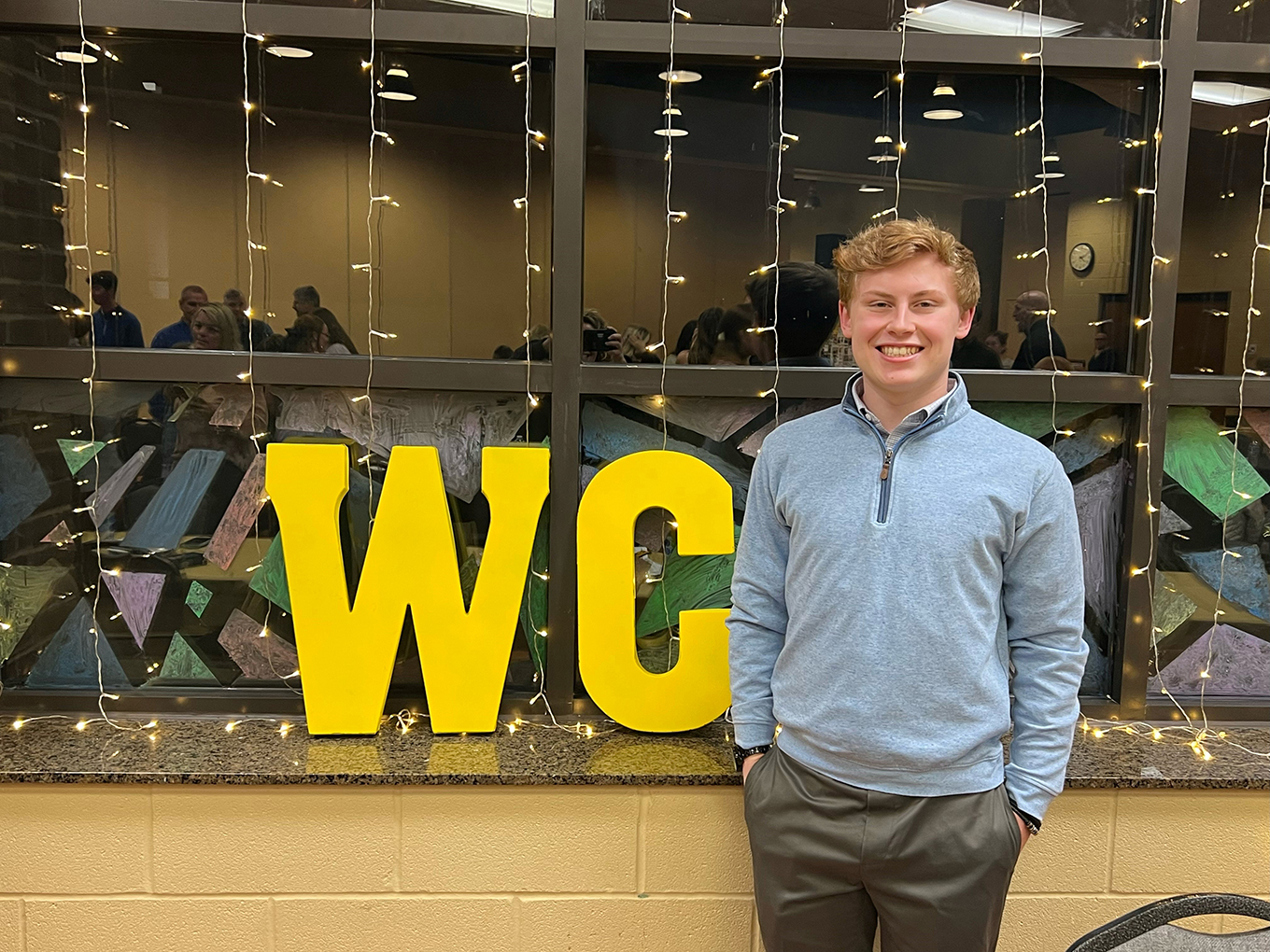 A person stands in front of a window with the letters "WC" behind him in gold.
