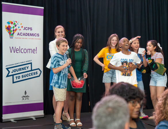 A group of kids stand on a podium with an adult, and one of the children is holding a mic while reaching into a bowl with red pieces of paper in it