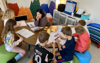 A teacher sits at a table surrounded by kids