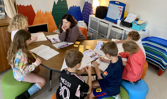 A teacher sits at a table surrounded by kids