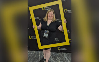 Heidi Givens poses while holding a gold frame similar to the National Geographic logo that adorns the background behind her