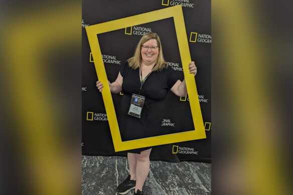 Heidi Givens poses while holding a gold frame similar to the National Geographic logo that adorns the background behind her