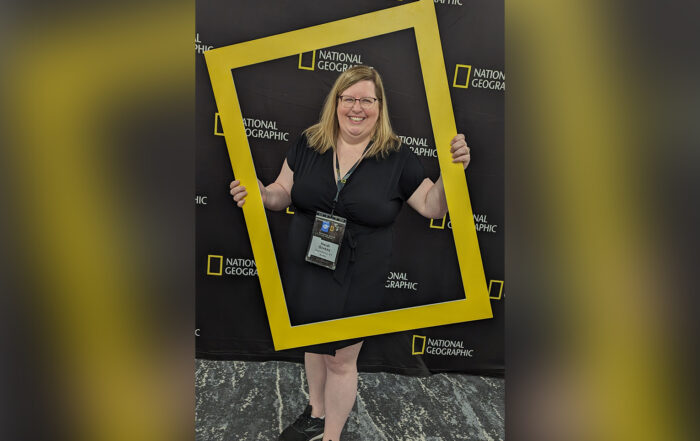 Heidi Givens poses while holding a gold frame similar to the National Geographic logo that adorns the background behind her