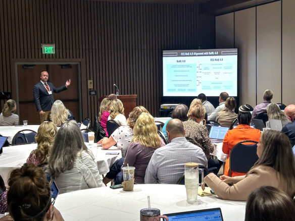 A room full of people watch a man present data on a screen