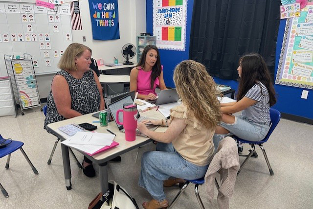 Four teachers sit at a table with papers and computers on it