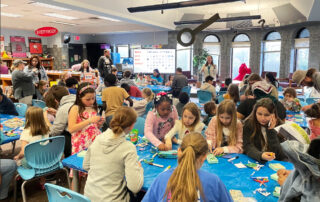 Groups of kids sit at tables in a classroom with multiple adults standing