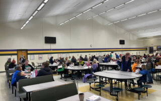 A group of people sit at various tables inside a gymnasium